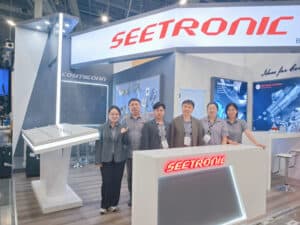A group stands in front of a "SEETRONIC" booth at a trade show, with promotional material and display items in the background.